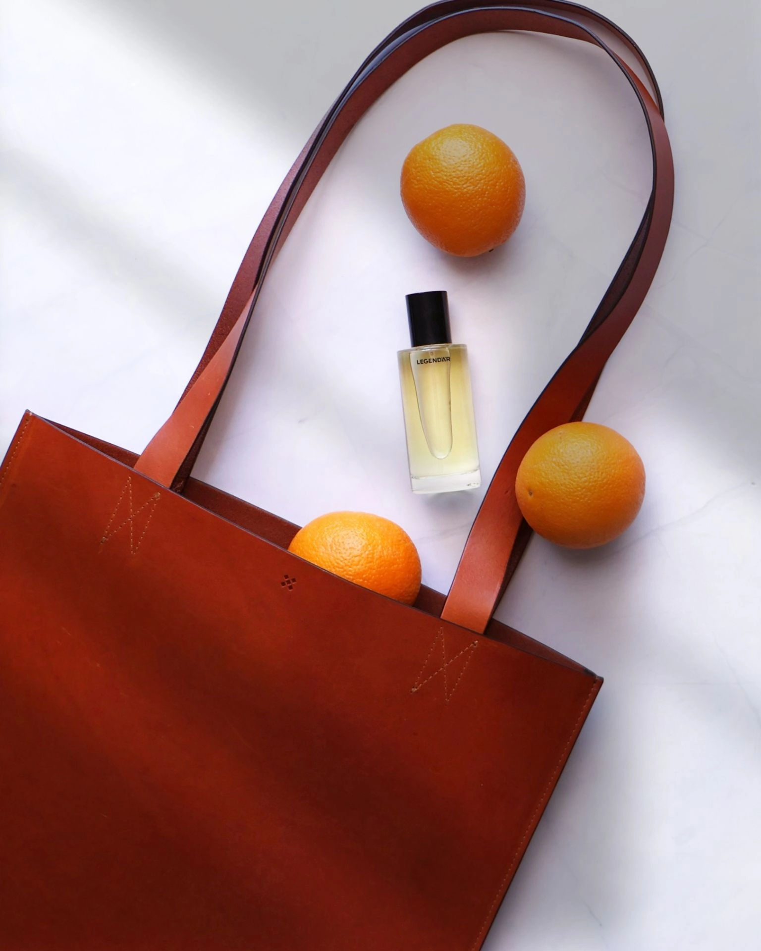 Legendär Tote Bag – Ledertragetasche mit Parfum und Orangen im Flatlay`` Legendär Tote Bag aus Leder mit Orangen und Eau de Parfum auf hellem Untergrund arrangiert